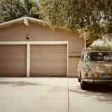 En beige veteranbil står parkerad framför ett garage med två portar. Kanske är det dags att bygga garage för att skydda den?