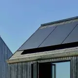 Tak med monokristallina solceller och träfasad mot blå himmel.