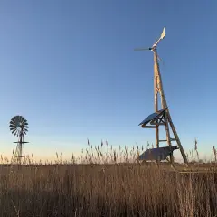 Två vindkraftverk, ett modernt med solpaneler och ett äldre, står i ett fält med torrt gräs. En vindturbin villa för hållbar energi.