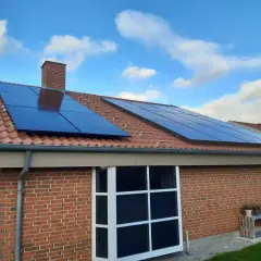 Solceller på ett tegeltak med blå himmel och moln. Ett hus med tegelväggar och stora fönster. Elcertifikat för solceller är en bra investering.