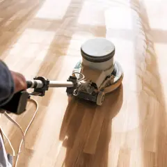 En person slipar parkettgolv med en maskin. Dammet virvlar upp och ljuset faller in från sidan.