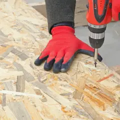 En hand med röd handske skruvar i en OSB-skiva. Tjockleken på OSB-skivan är tydlig när skruven dras åt.