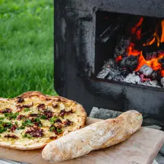 En pizza och ett bröd ligger på en skärbräda bredvid en pizzaugn med glödande ved.