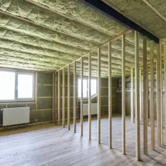 Rummet är under renovering med träreglar och ljudisolering på väggar och tak. Två fönster med vita element under.