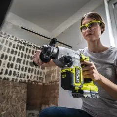 Kvinna med skyddsglasögon använder en slagborrmaskin för att borra i tegelvägg. Damm virvlar upp.