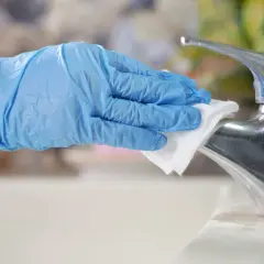 A gloved hand wipes a chrome faucet with a white cloth.