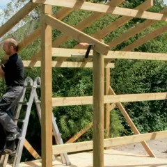 Man on ladder building wooden shed frame with drill.