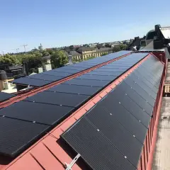 Rows of black solar panels installed on a red metal roof with buildings and trees in the background.