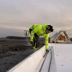 Arbetare i varselkläder förbereder armering för betongsten grund. Takstolar syns i bakgrunden.