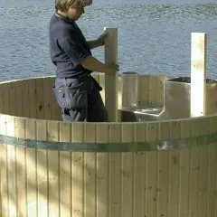 Man bygger badtunna vid en sjö. Träet är ljust och en metallring håller ihop tunnan.