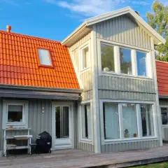 Grått trähus med orange takpannor. En person klättrar på en stege, kanske för att måla takpannor eller utföra annat underhåll.