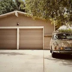 En beige veteranbil står parkerad framför ett garage med två portar. Kanske är det dags att bygga garage för att skydda den?