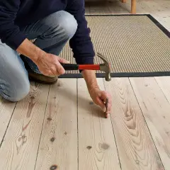 En person knäböjer och slår en spik i ett trägolv för att åtgärda ett knarrande golv.