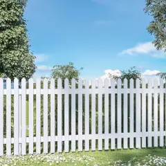 En vit staket med spetsiga plank stående ribbor avgränsar en grönskande trädgård under en blå himmel med moln.