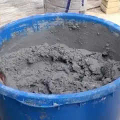 En blå balja fylld med grå cement som någon håller på att blanda. Tegelstenar syns i bakgrunden.
