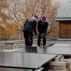 Två personer installerar solceller på ett platt tak. Höstfärger syns i bakgrunden.