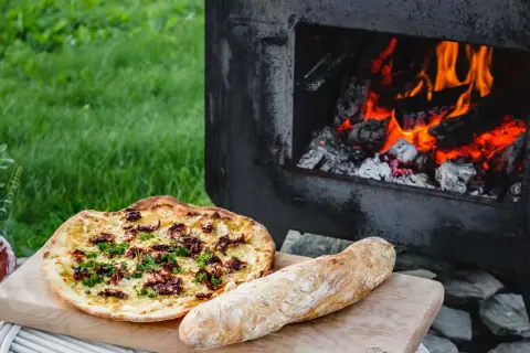 En pizza och ett bröd ligger på en skärbräda bredvid en pizzaugn med glödande ved.