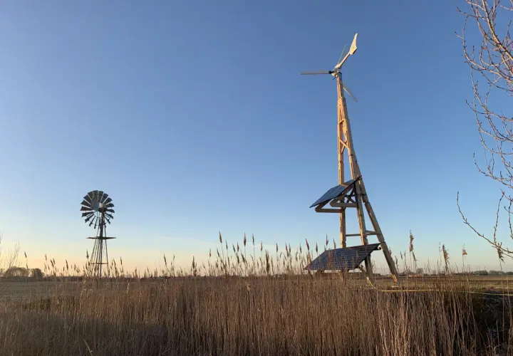 Två vindkraftverk, ett modernt med solpaneler och ett äldre, står i ett fält med torrt gräs. En vindturbin villa för hållbar energi.