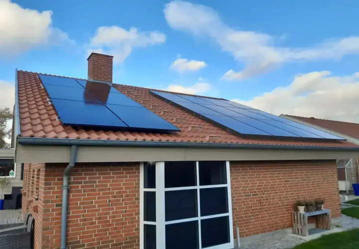 Solceller på ett tegeltak med blå himmel och moln. Ett hus med tegelväggar och stora fönster. Elcertifikat för solceller är en bra investering.