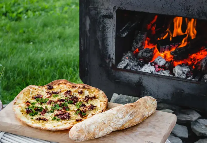 En pizza och ett bröd ligger på en skärbräda bredvid en pizzaugn med glödande ved.