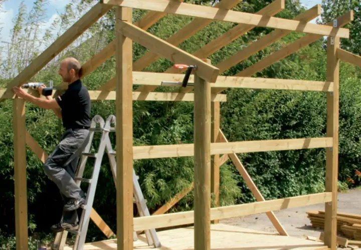 Man on ladder building wooden shed frame with drill.