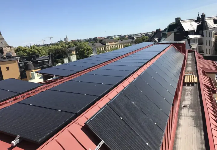 Rows of black solar panels installed on a red metal roof with buildings and trees in the background.