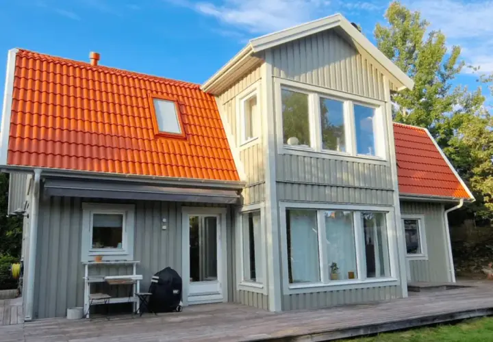 Grått trähus med orange takpannor. En person klättrar på en stege, kanske för att måla takpannor eller utföra annat underhåll.
