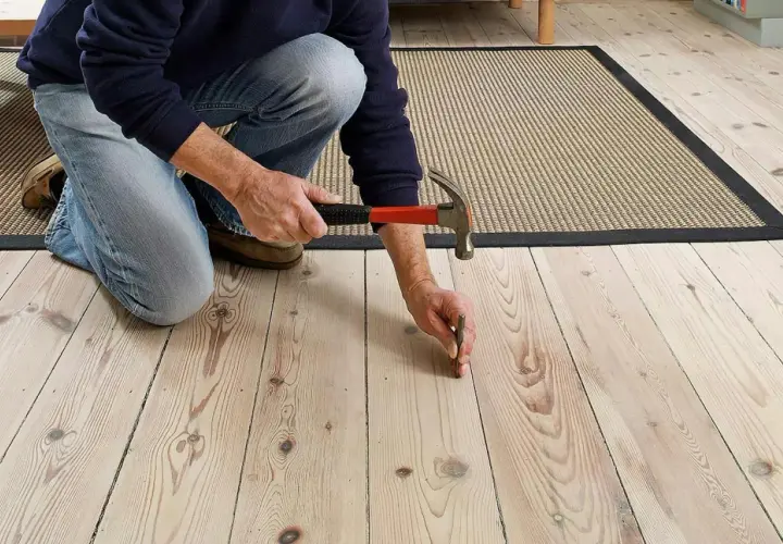 En person knäböjer och slår en spik i ett trägolv för att åtgärda ett knarrande golv.