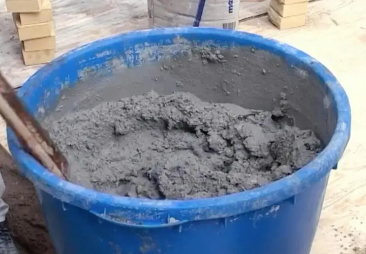En blå balja fylld med grå cement som någon håller på att blanda. Tegelstenar syns i bakgrunden.