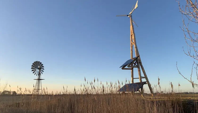 Två vindkraftverk, ett modernt med solpaneler och ett äldre, står i ett fält med torrt gräs. En vindturbin villa för hållbar energi.