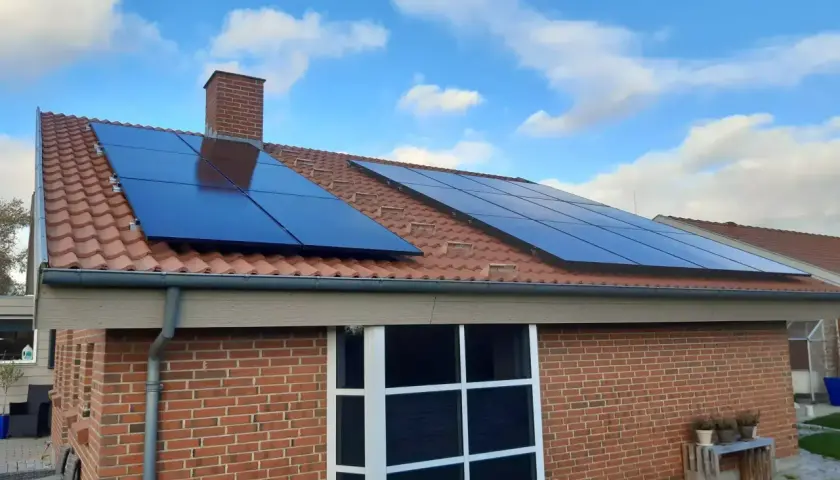 Solceller på ett tegeltak med blå himmel och moln. Ett hus med tegelväggar och stora fönster. Elcertifikat för solceller är en bra investering.