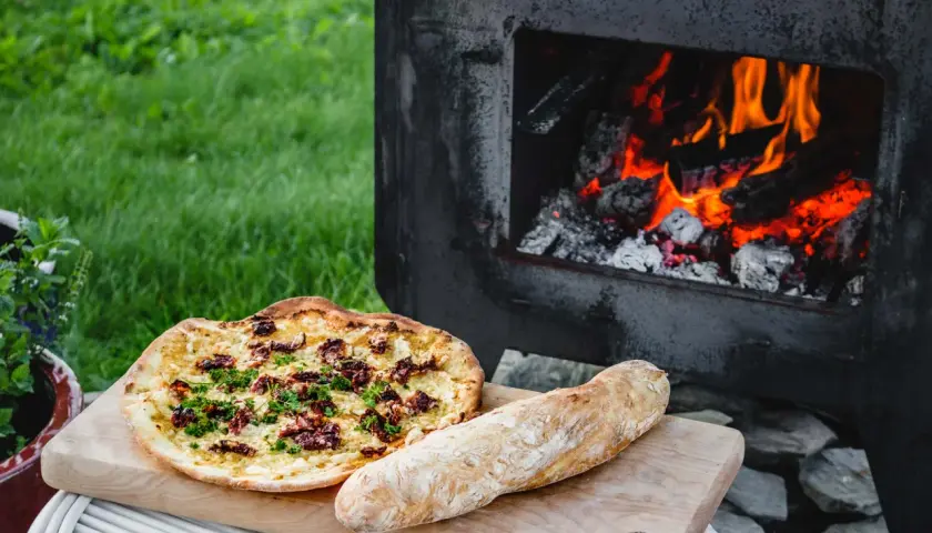 En pizza och ett bröd ligger på en skärbräda bredvid en pizzaugn med glödande ved.