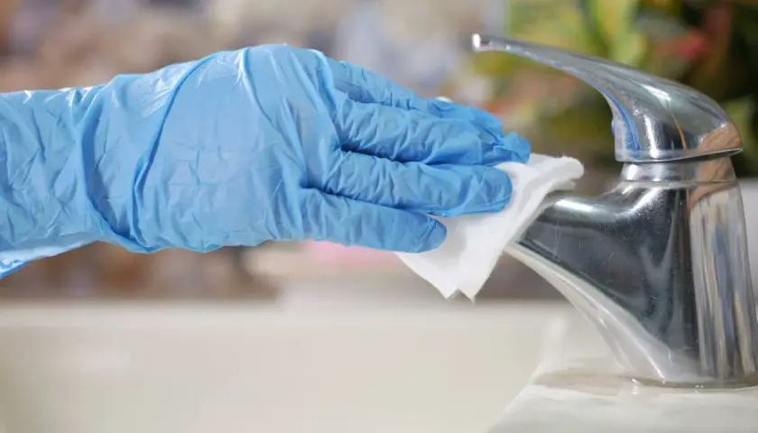 A gloved hand wipes a chrome faucet with a white cloth.