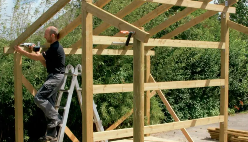 Man on ladder building wooden shed frame with drill.