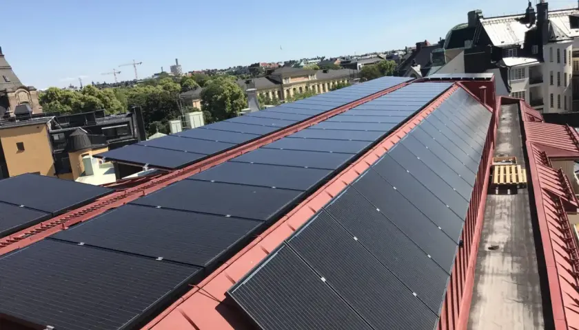 Rows of black solar panels installed on a red metal roof with buildings and trees in the background.