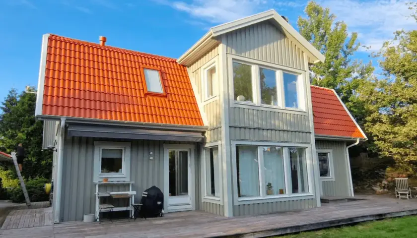 Grått trähus med orange takpannor. En person klättrar på en stege, kanske för att måla takpannor eller utföra annat underhåll.