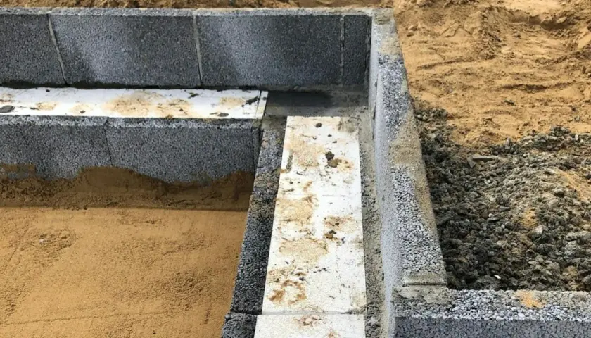 Grundläggande arbete med lecablock. En del av grunden är färdigbyggd med grå lecablock och isolering. Sand och jord syns runt omkring.