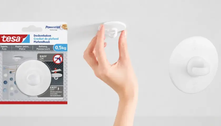En hand fäster en vit krok i taket. Tesa Powerstrips-krok för tak, enkel att sätta upp och ta ner.