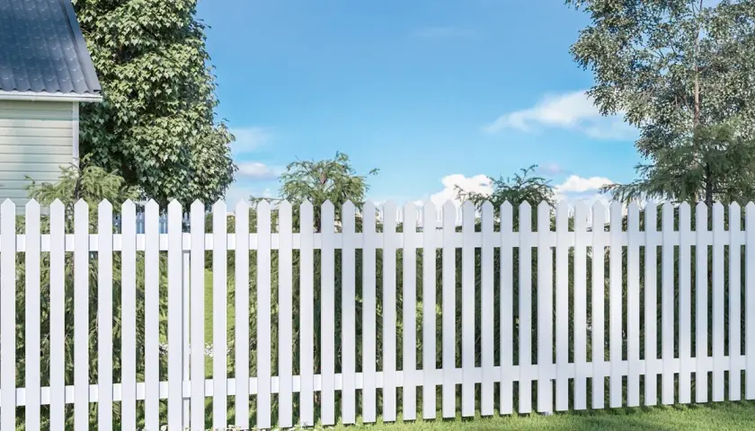 En vit staket med spetsiga plank stående ribbor avgränsar en grönskande trädgård under en blå himmel med moln.