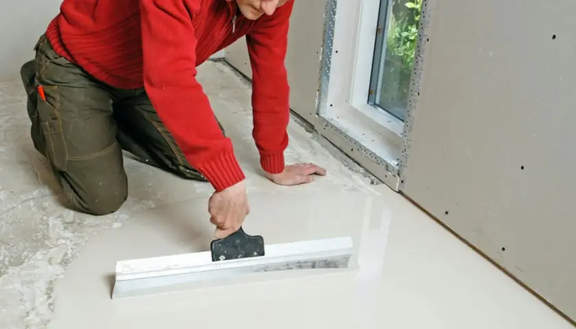 Man i röd tröja och gröna byxor jämnar ut flytspackel för att gjuta betonggolv.