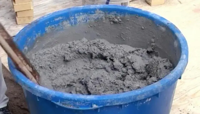 En blå balja fylld med grå cement som någon håller på att blanda. Tegelstenar syns i bakgrunden.