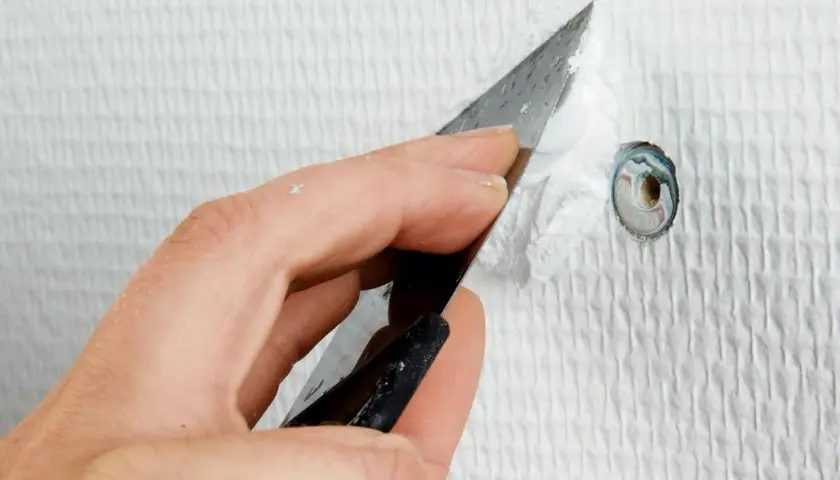 Hand spacklar ett hål i en vit, strukturerad vägg med en spackel.