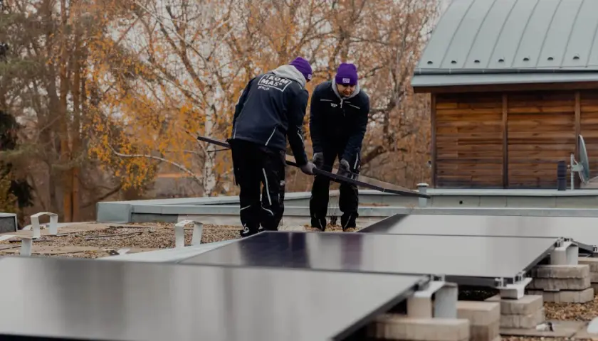 Två personer installerar solceller på ett platt tak. Höstfärger syns i bakgrunden.