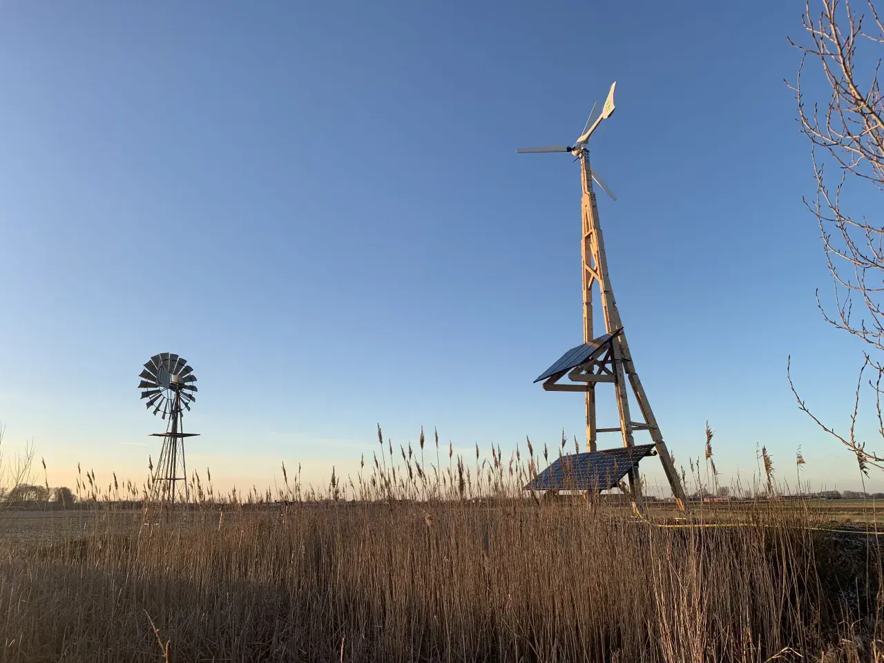 Två vindkraftverk, ett modernt med solpaneler och ett äldre, står i ett fält med torrt gräs. En vindturbin villa för hållbar energi.