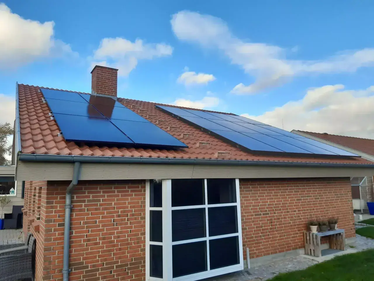 Solceller på ett tegeltak med blå himmel och moln. Ett hus med tegelväggar och stora fönster. Elcertifikat för solceller är en bra investering.