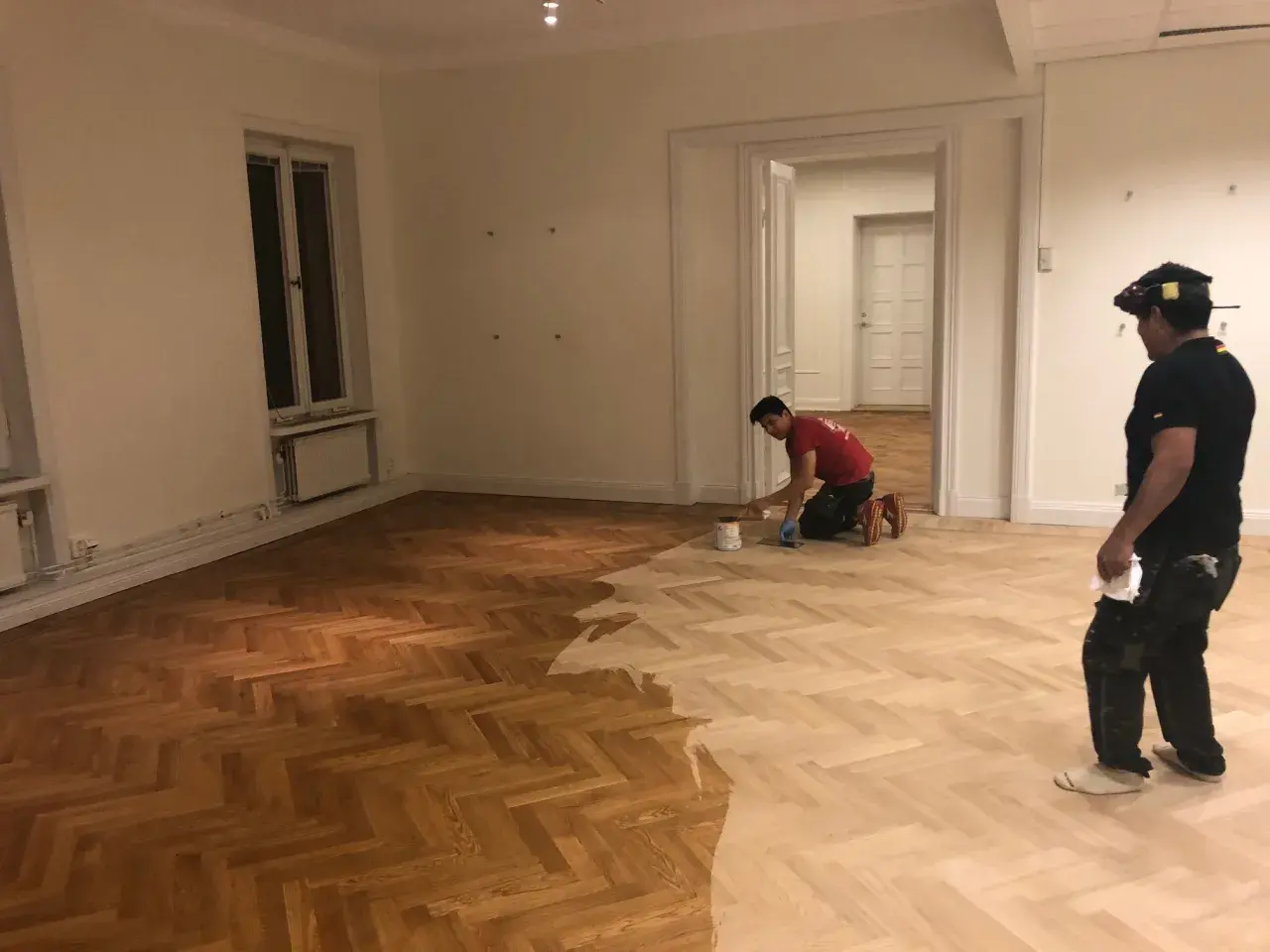 Två män arbetar med att slipa fiskbensparkett i ett rum. Golvet är halvt behandlat, med en mörkare, slipad del och en ljusare, obehandlad del.
