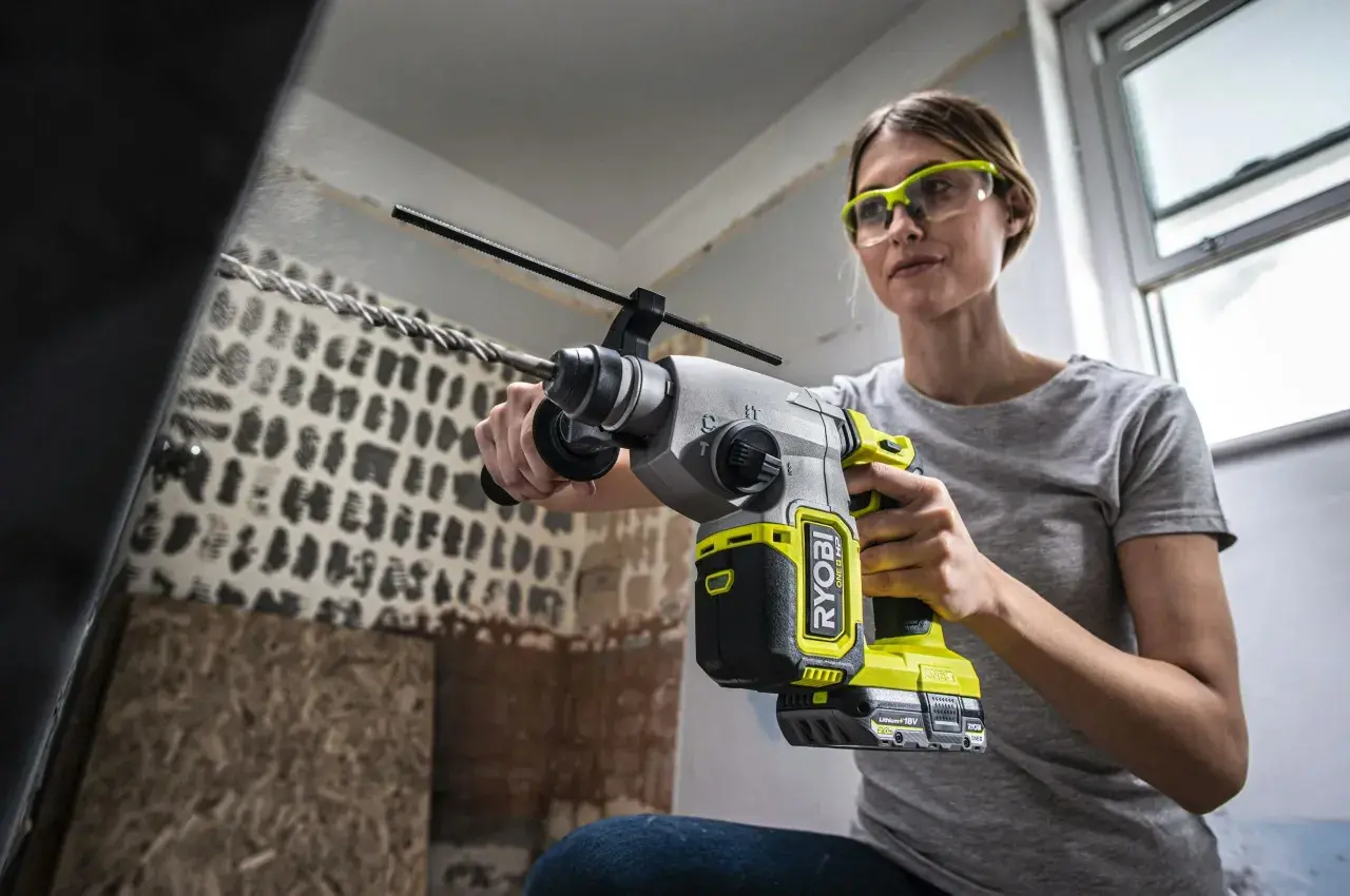 Kvinna med skyddsglasögon använder en slagborrmaskin för att borra i tegelvägg. Damm virvlar upp.