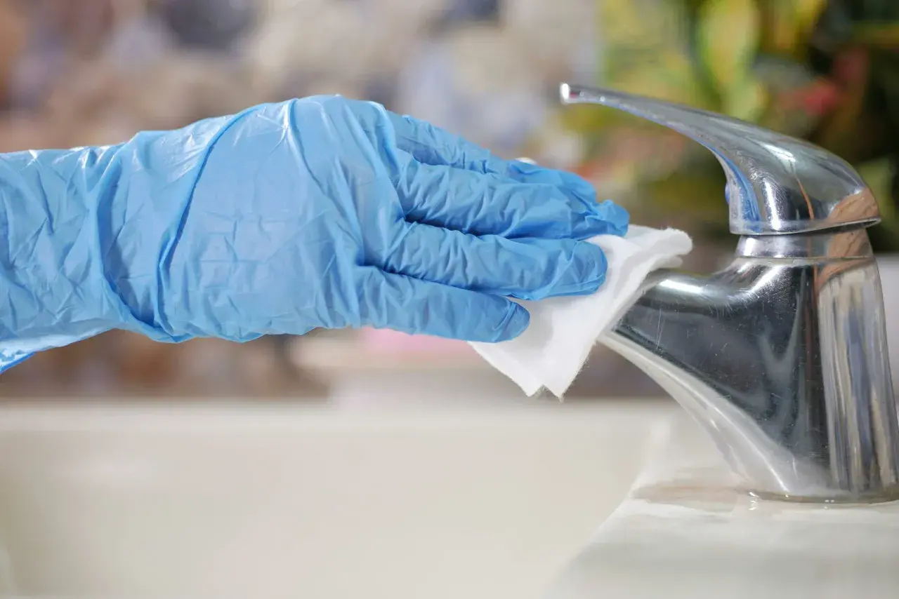 A gloved hand wipes a chrome faucet with a white cloth.