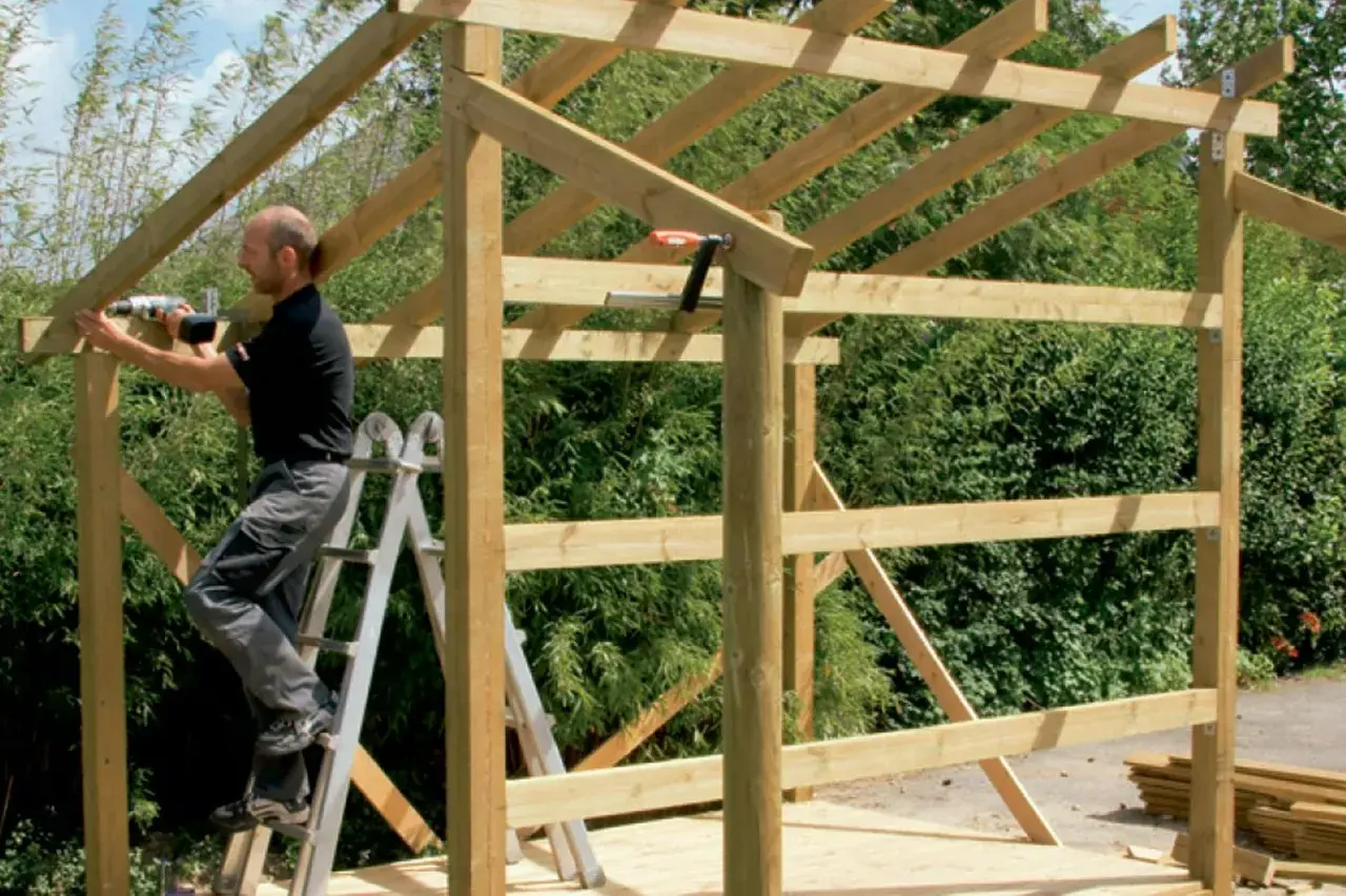 Man on ladder building wooden shed frame with drill.