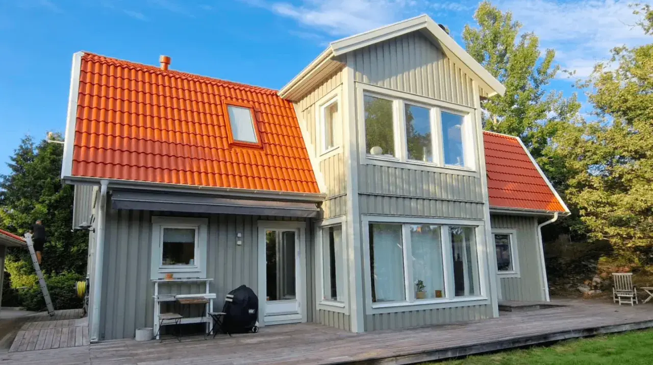 Grått trähus med orange takpannor. En person klättrar på en stege, kanske för att måla takpannor eller utföra annat underhåll.